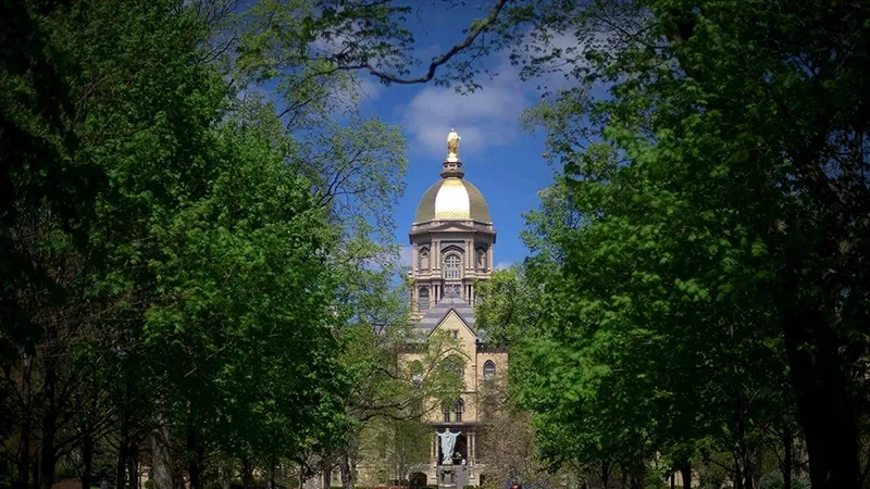 The Golden Dome atop the Main Building at the University of Notre Dame is seen through a frame of lush green trees on a bright spring day. A statue of Jesus with outstretched arms stands in front of the Main Building.